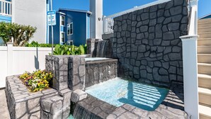 Une fontaine en pierre avec un petit bassin, entourée d’une clôture blanche et d’un bâtiment bleu.