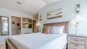 Une chambre à coucher avec un grand lit, des tables de chevet, une commode en bois, un tableau au mur et une porte donnant sur une autre pièce.