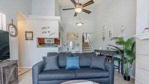 Un salon avec un canapé gris, une table basse avec des télécommandes, un ventilateur de plafond et, au fond, une cuisine.
