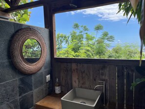A bathroom with a stone wall, a woven mirror, a sink, and a view of greenery. - Villa La Pointe. 3 bedrooms. Swimming pool and ocean view. 600 meters from the Saline Lagoon. (Saline les bains)
