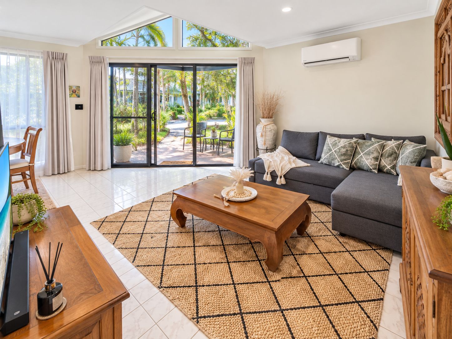 A modern living room with a sofa, coffee table, and a view of a tropical garden through glass doors.
