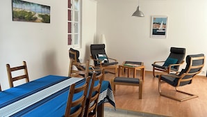A dining area with a long table, wooden chairs, and a blue tablecloth. There are two lounge chairs and a small table with a lamp. The room has white walls and a tiled floor. - MATOUNE pleasant, 4/6 person house near Lake Leon  (Léon)