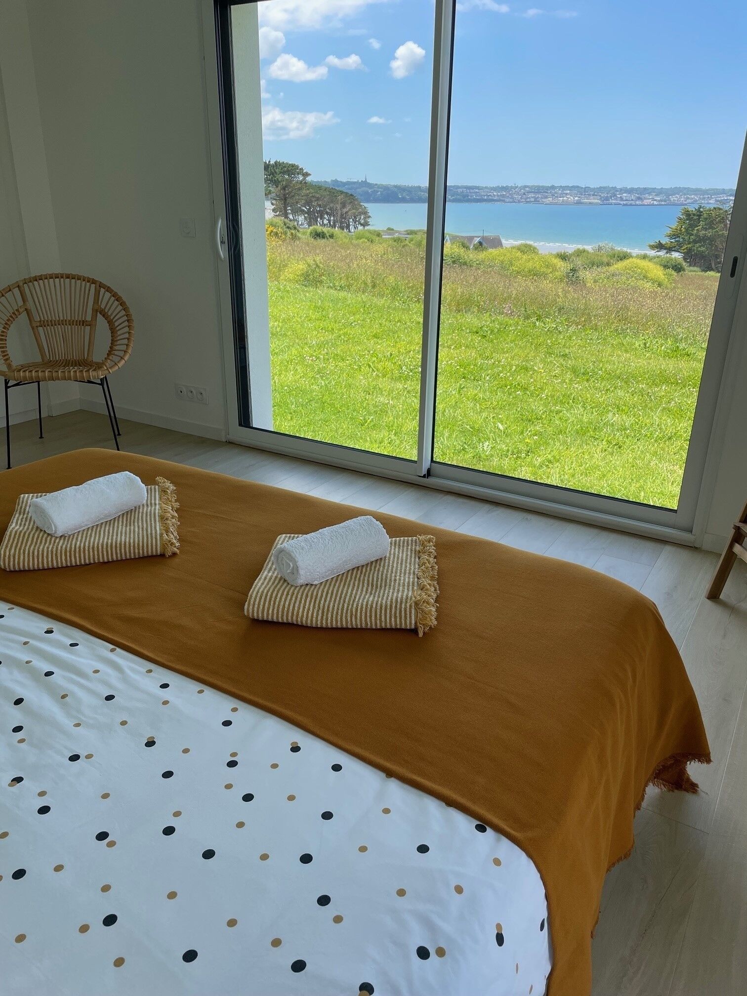 Een slaapkamer met een groot bed, twee handdoeken, een stoel en uitzicht op een grasveld en water door een groot raam.
