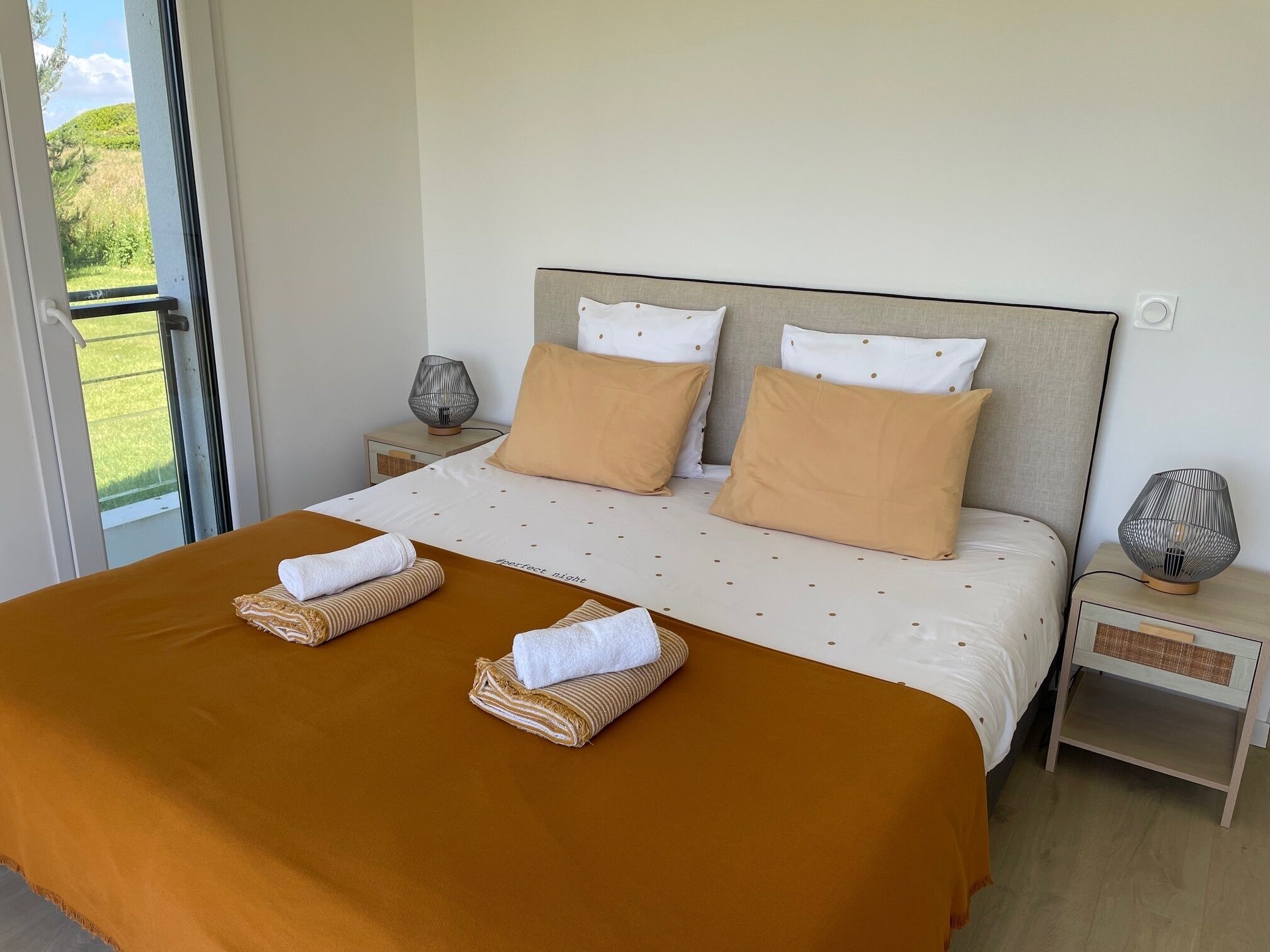 Une chambre d’hôtel avec un grand lit, deux lampes de chevet, une vue sur la verdure grâce à une porte coulissante, et un lit parfaitement fait avec des serviettes.