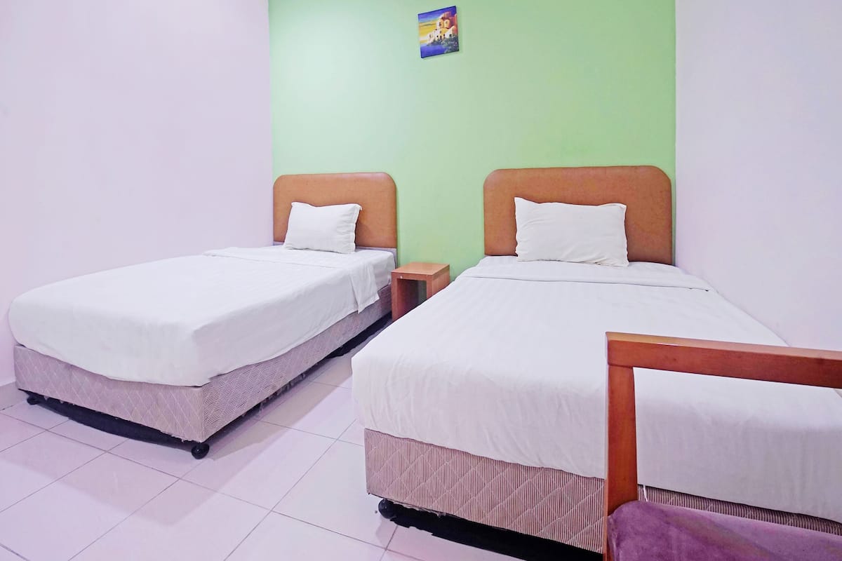 A room with two beds, each with a wooden headboard and white bedding. There is a small table and a nightstand next to each bed. The walls are painted light green, and there is a small picture on the wall above the beds.