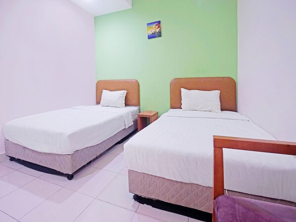 Habitación con dos camas, cada una con cabecero de madera y ropa de cama blanca. Hay una mesita y una mesita de noche junto a cada cama. Las paredes están pintadas de verde claro, y hay un cuadro pequeño en la pared sobre las camas.