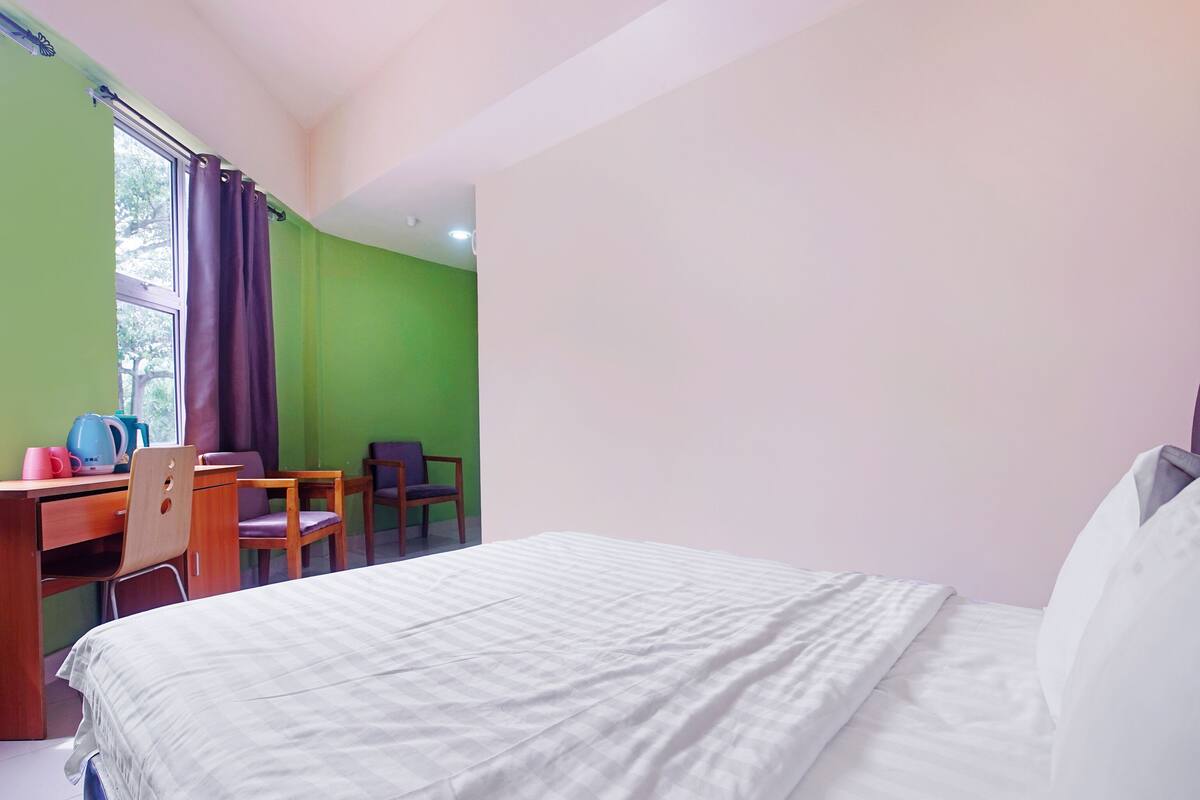 A hotel room with a bed, a desk with chairs, a window with curtains, and a green wall.