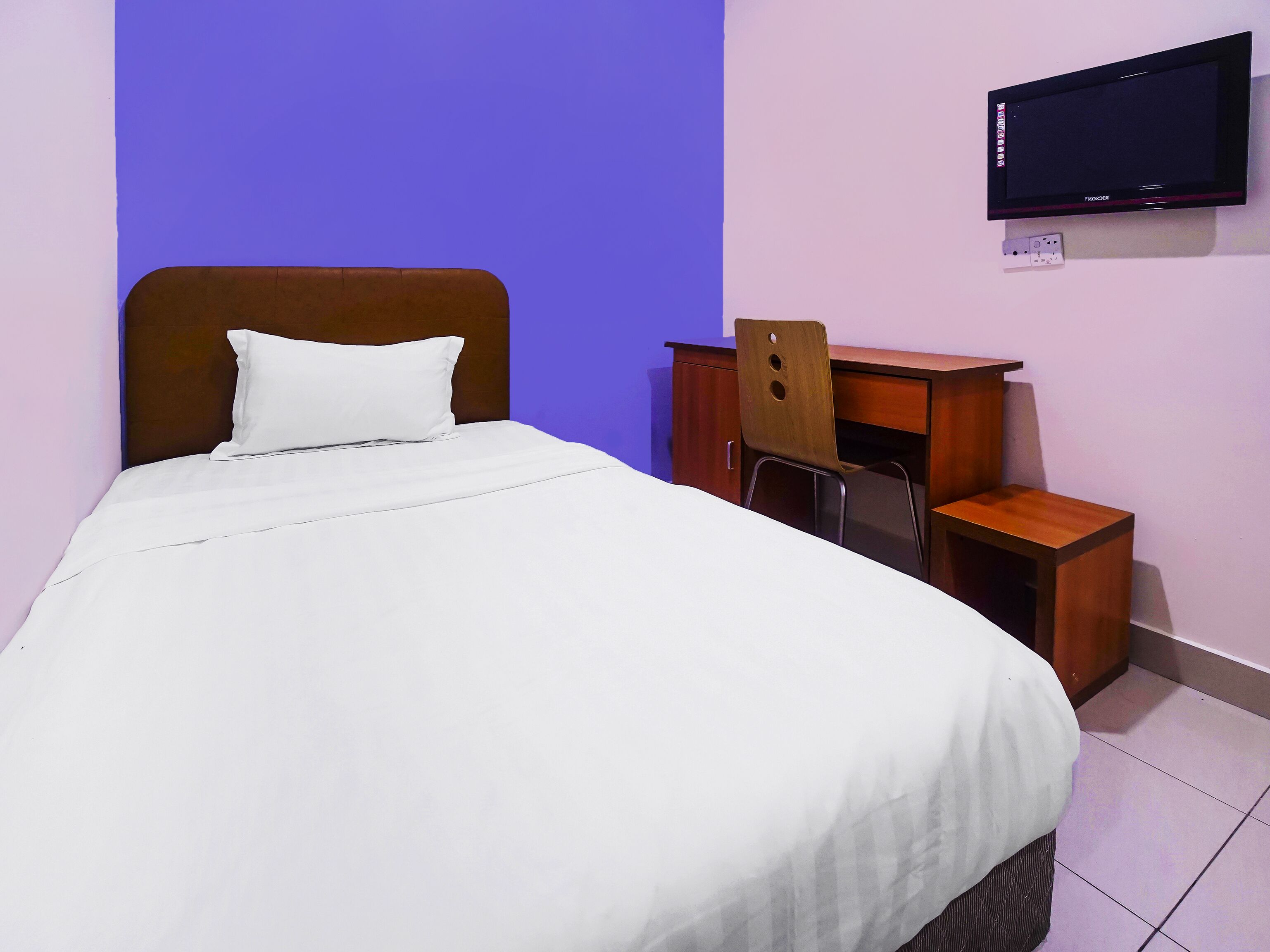A neatly made bed with white bedding, a wooden headboard, and a small wooden desk with a chair. A flat-screen TV is mounted on the wall above the desk.