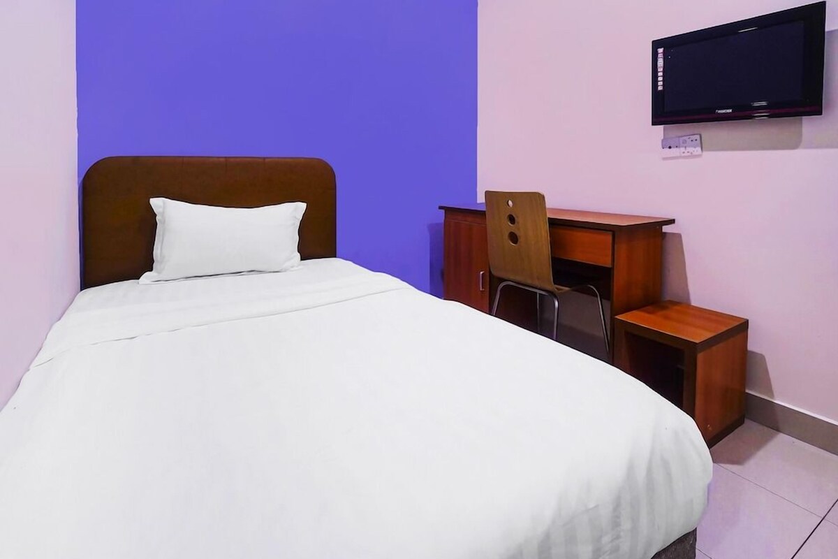 A bed with a white mattress and a wooden headboard, a wooden desk with a chair, and a television mounted on the wall.