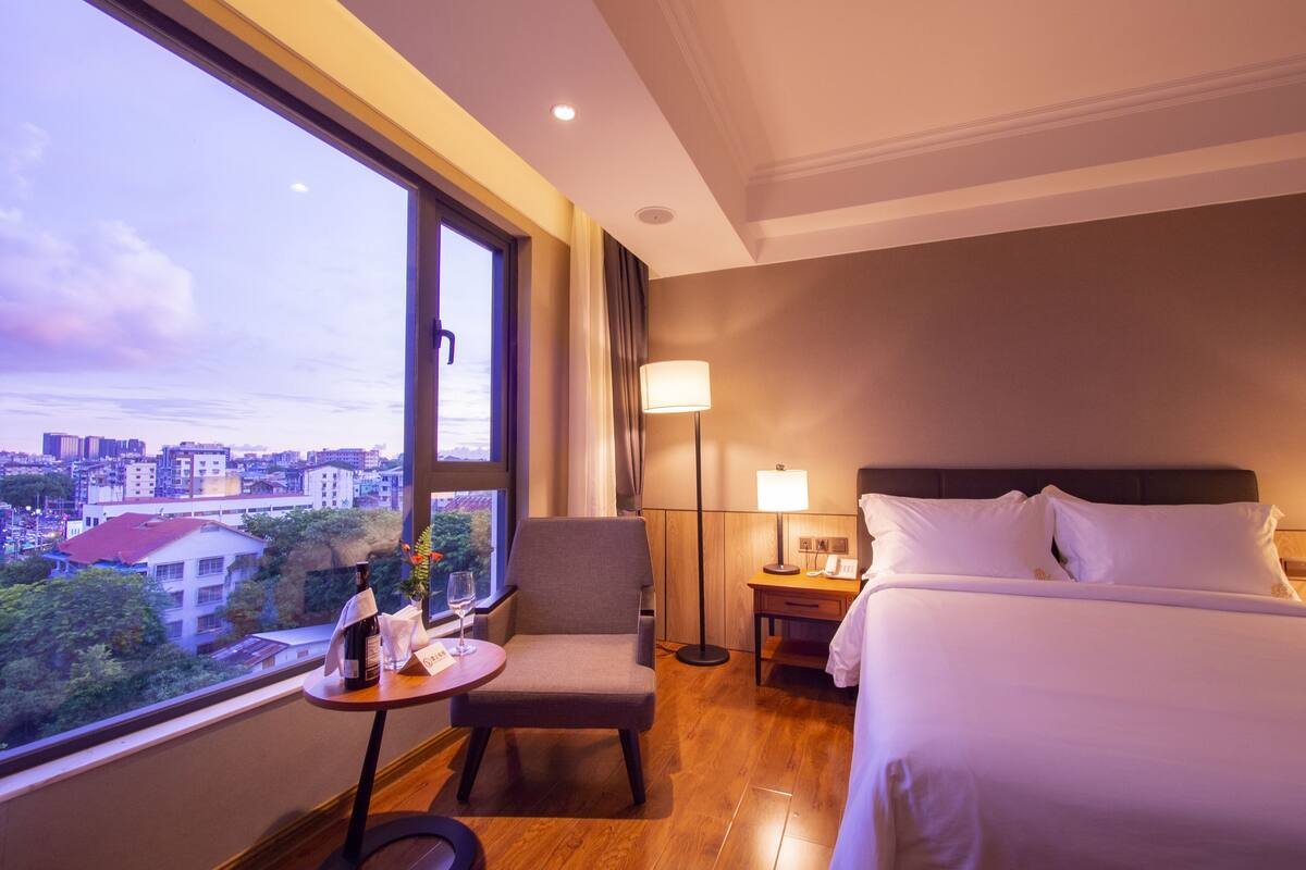 A hotel room with a large window, a bed, a chair, a small table with a bottle and glass, and a nightstand with a lamp.