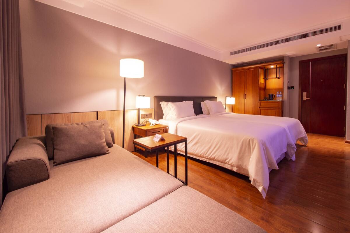 A hotel room with a large bed, a smaller sofa, a bedside table with a lamp, a nightstand, and a wooden wardrobe.