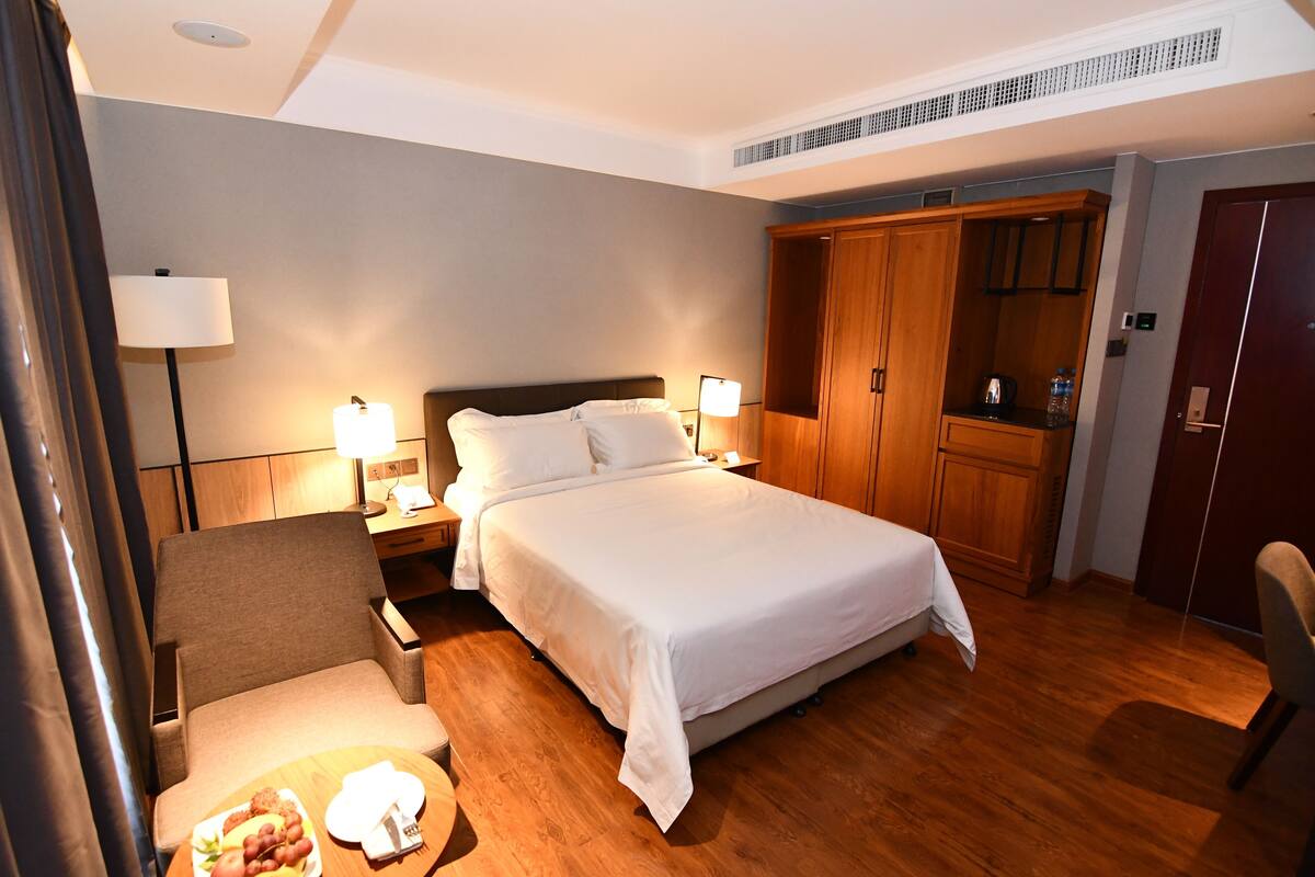 A hotel room with a bed, two bedside tables with lamps, a wooden wardrobe, a chair, and a small table with a plate of food.