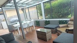 Ein überdachter Patio-Bereich mit Esstisch und Stühlen, Korbmöbeln und Blick auf die umliegende Bepflanzung.
