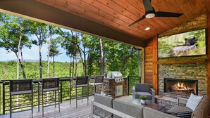 Eine Terrasse im Freien mit Kamin aus Stein, Grill und Sitzgelegenheit mit Blick auf eine bewaldete Landschaft.