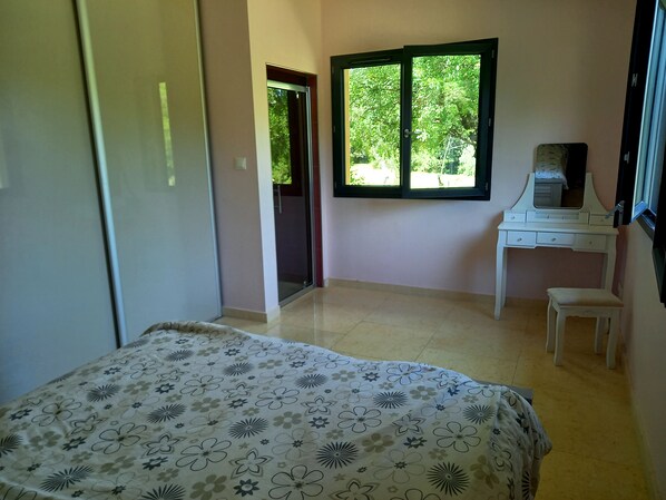 A bedroom with a bed, a desk, a chair, and a window with a view of greenery.