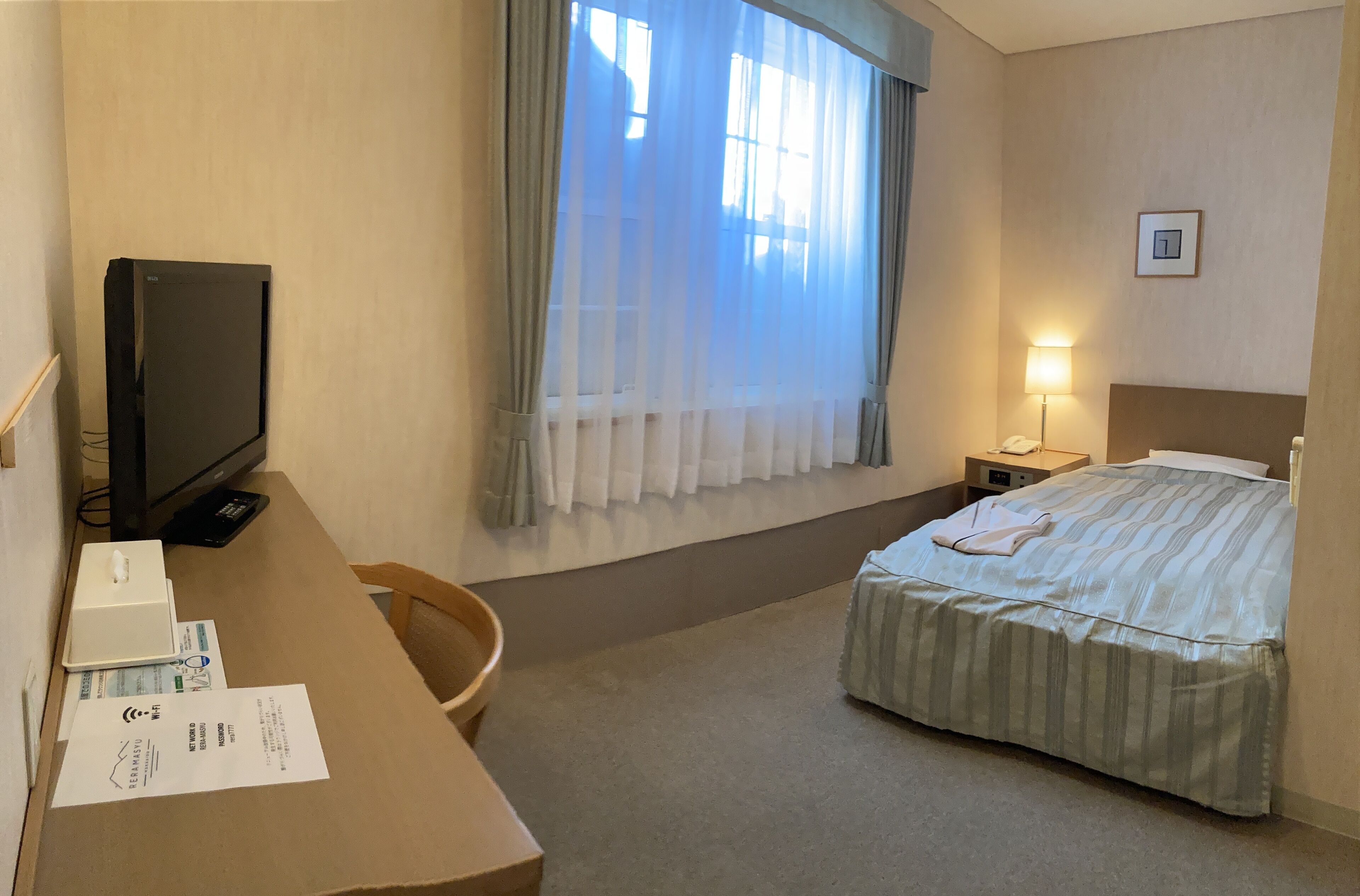 Una habitación de hotel con cama, un escritorio con computadora y papeles, un televisor y una ventana con cortinas.