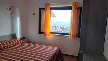A hotel room with a bed, a window with curtains, and a view of the sea and buildings.