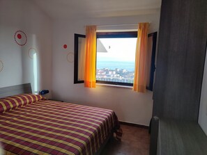 A hotel room with a bed, a window with curtains, and a view of the sea and buildings. - Peaceful Sea -View Apartment in Falerna |Pool, Tennis & Beach Nearby (Calabria)