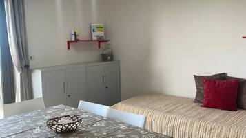A dining area with a table set for two, a grey cabinet, and a bed with a striped blanket and pillows.
