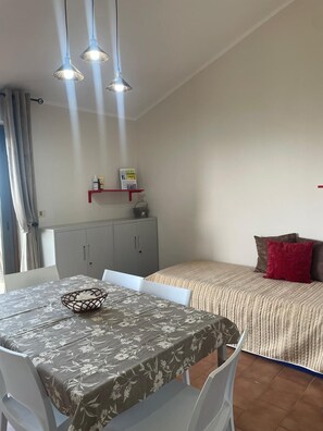 A dining area with a table set for two, a grey cabinet, and a bed with a striped blanket and pillows. - Peaceful Sea -View Apartment in Falerna |Pool, Tennis & Beach Nearby (Calabria)
