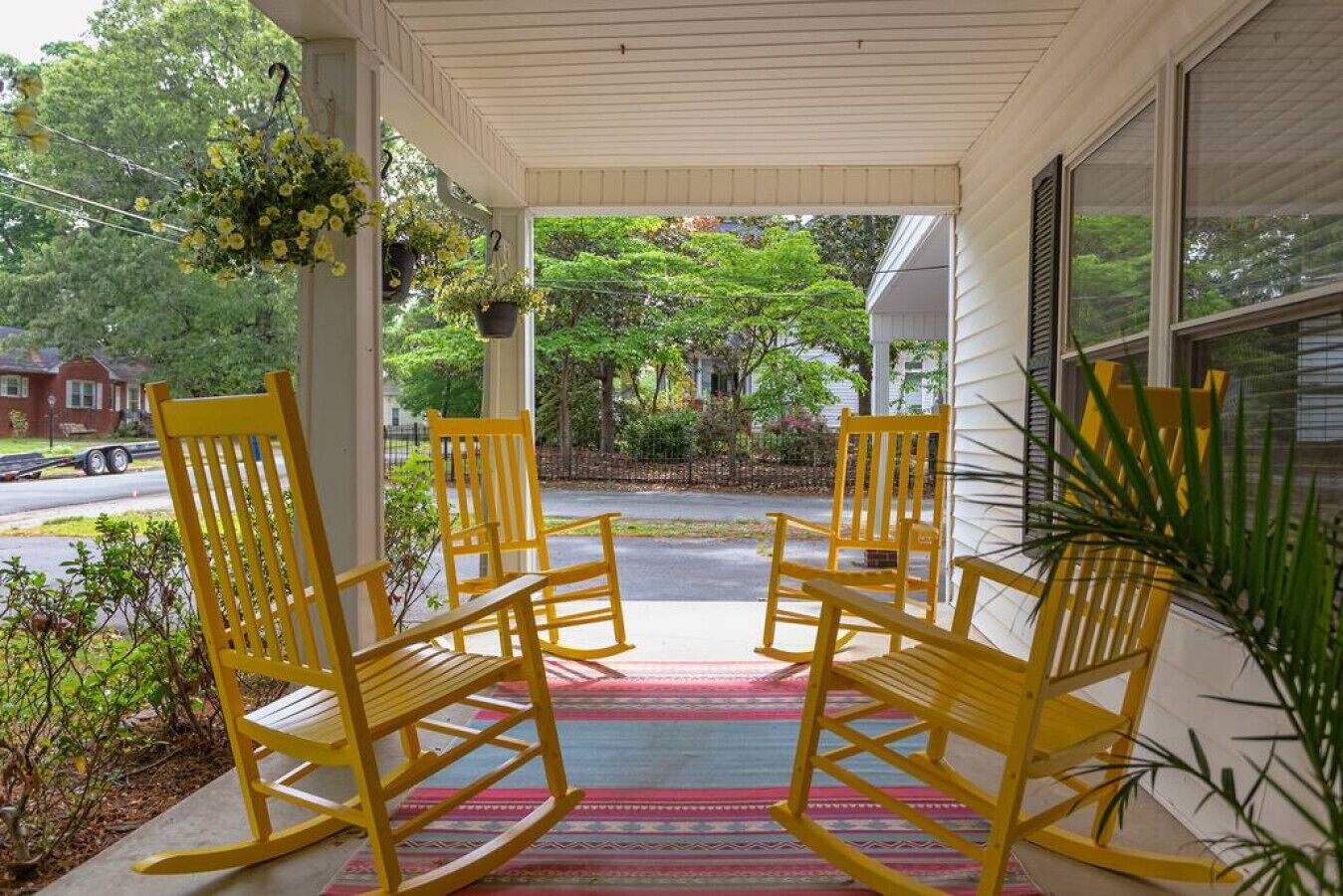 A porch with four yellow rocking chairs, a colorful rug, and hanging flower baskets.