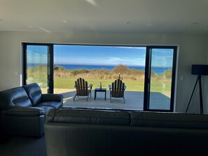 A modern living room with a large sliding glass door leading to a deck with two Adirondack chairs and a small table, overlooking a grassy area and the ocean.