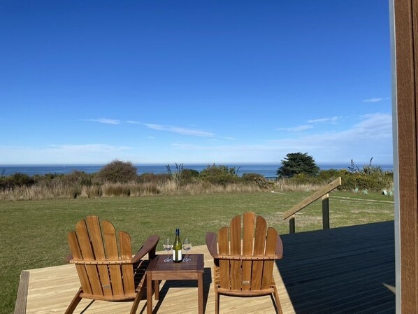Two wooden Adirondack chairs on a wooden deck with a view of the ocean.