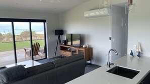 Una cocina moderna con isla integrada, un comedor con sofá y televisor, y un amplio ventanal corredero que da a una terraza con vista al océano.