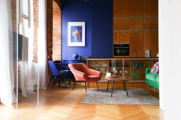 A modern living room with a herringbone wooden floor, a red armchair, a green sofa, and a glass cabinet with kitchenware.
