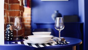 Ein Esstisch mit weißen Tellern, einem Weinglas und Besteck. Auf dem Tisch stehen eine schwarze, gepunktete Vase und eine kleine Figur. Der Raum hat blaue Wände und eine Ziegelwand im Hintergrund.
