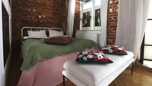 Ein Schlafzimmer mit einer Ziegelwand, einem Bett mit einer grünen und pinkfarbenen Bettdecke, weißen Kissen und einer Bank mit blumigen Kissen.