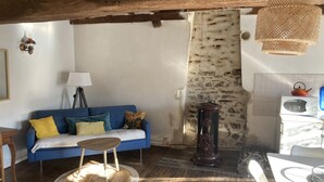 Ein gemütliches Wohnzimmer mit einem blauen Sofa, einem kleinen runden Tisch und einer Lampe. Eine rustikale Steinwand und eine Küchenzeile mit Mikrowelle und Wasserkocher sind ebenfalls vorhanden.