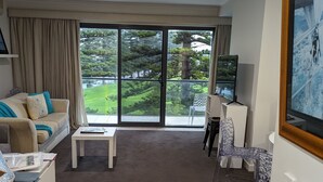 Een moderne woonkamer met een bank, salontafel en transparante stoelen. Een televisie staat op een dressoir en er is een groot raam met uitzicht op groen.