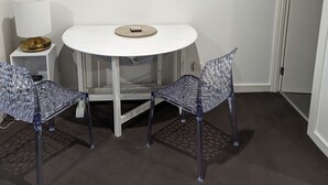 Een kleine, moderne kamer met een witte tafel en twee stoelen, een lamp en twee ingekaderde foto's aan de muur.
