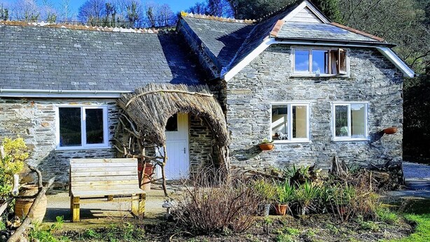 A stone house with a thatched roof entrance, a wooden bench outside, and a garden with potted plants. - A cosy riverside cottage in the Devon countryside on the Edge of Dartmoor. (Yelverton)