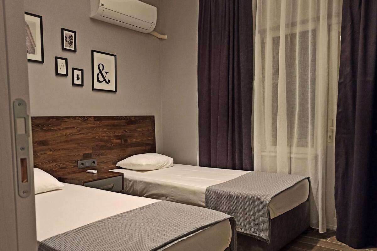 A hotel room with two beds, a wooden headboard, a wall-mounted air conditioner, and a window with dark curtains.