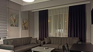 A modern living room with a grey sofa, a small coffee table, and a dining table with chairs. There are two paintings on the wall and a large window with dark curtains. - Merve Apart Otel (Alanya)