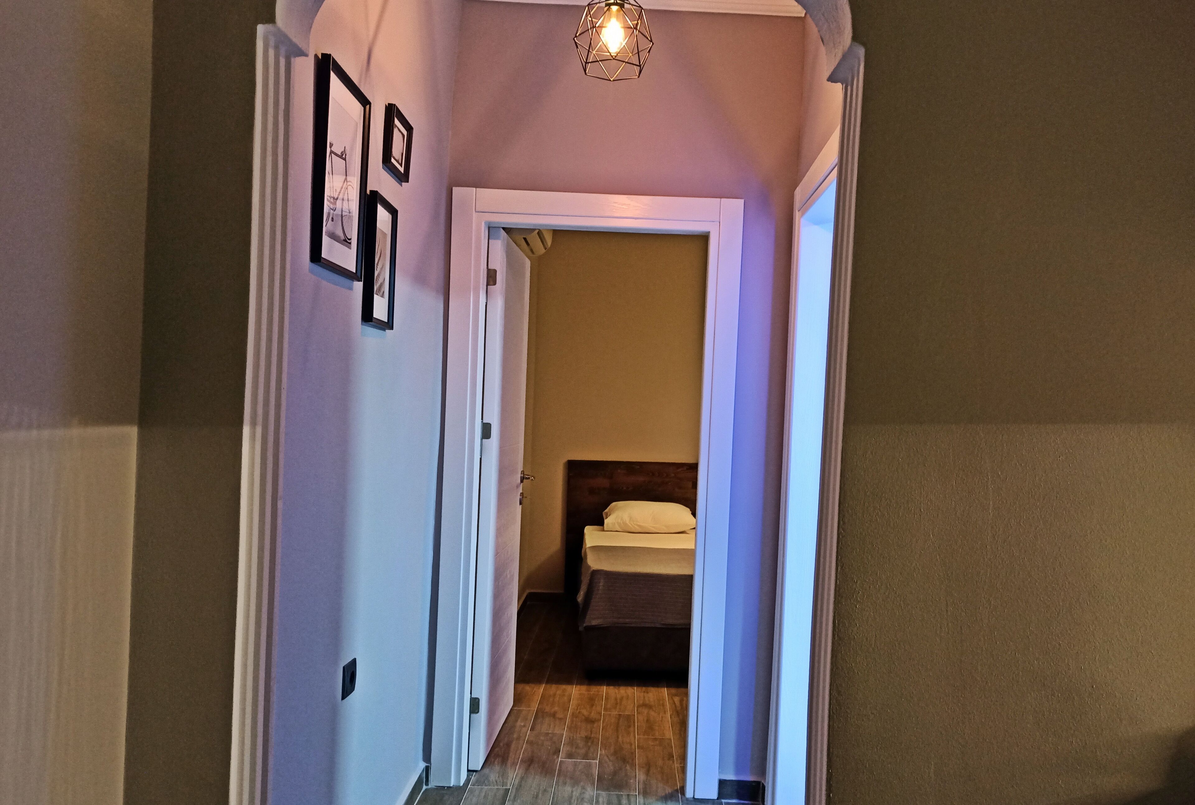 Um corredor estreito com uma cama no fim, emoldurado por paredes brancas e um aparelho de iluminação acima.