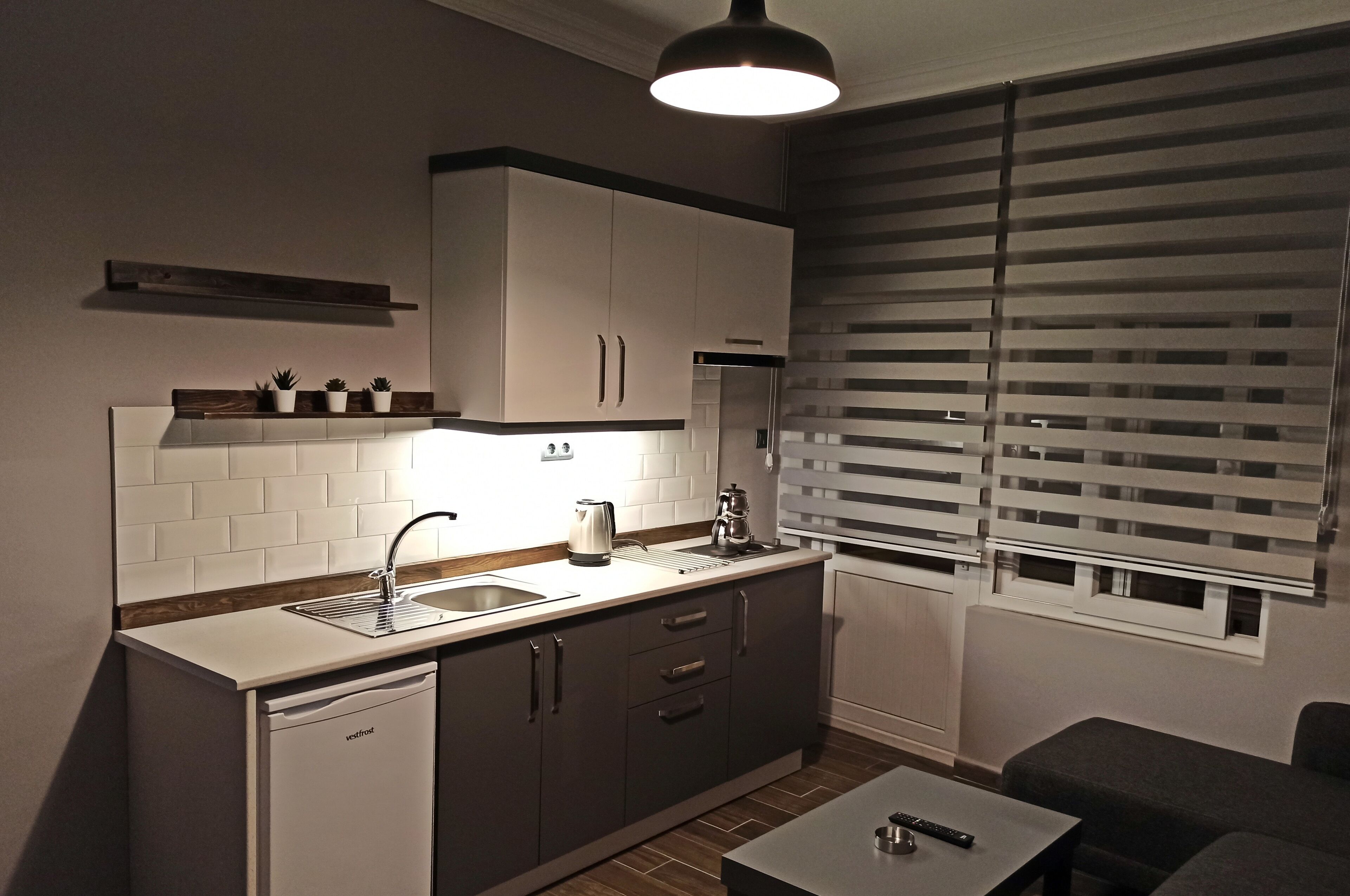 A compact kitchen with a dishwasher, a sink, and a coffee maker on the counter. There is a window with blinds and a small shelf above the counter.
