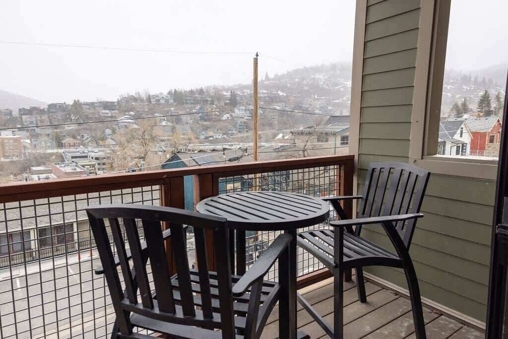 Un balcon avec deux chaises et une table, donnant sur un quartier résidentiel avec des maisons et un paysage vallonné.