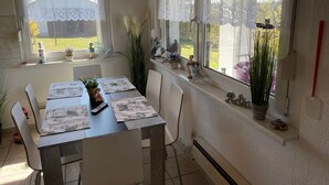 A dining area with a table set for two, two windows with lace curtains, and various decorative items on the windowsills. - Hobbithaus resort (Eggesin)