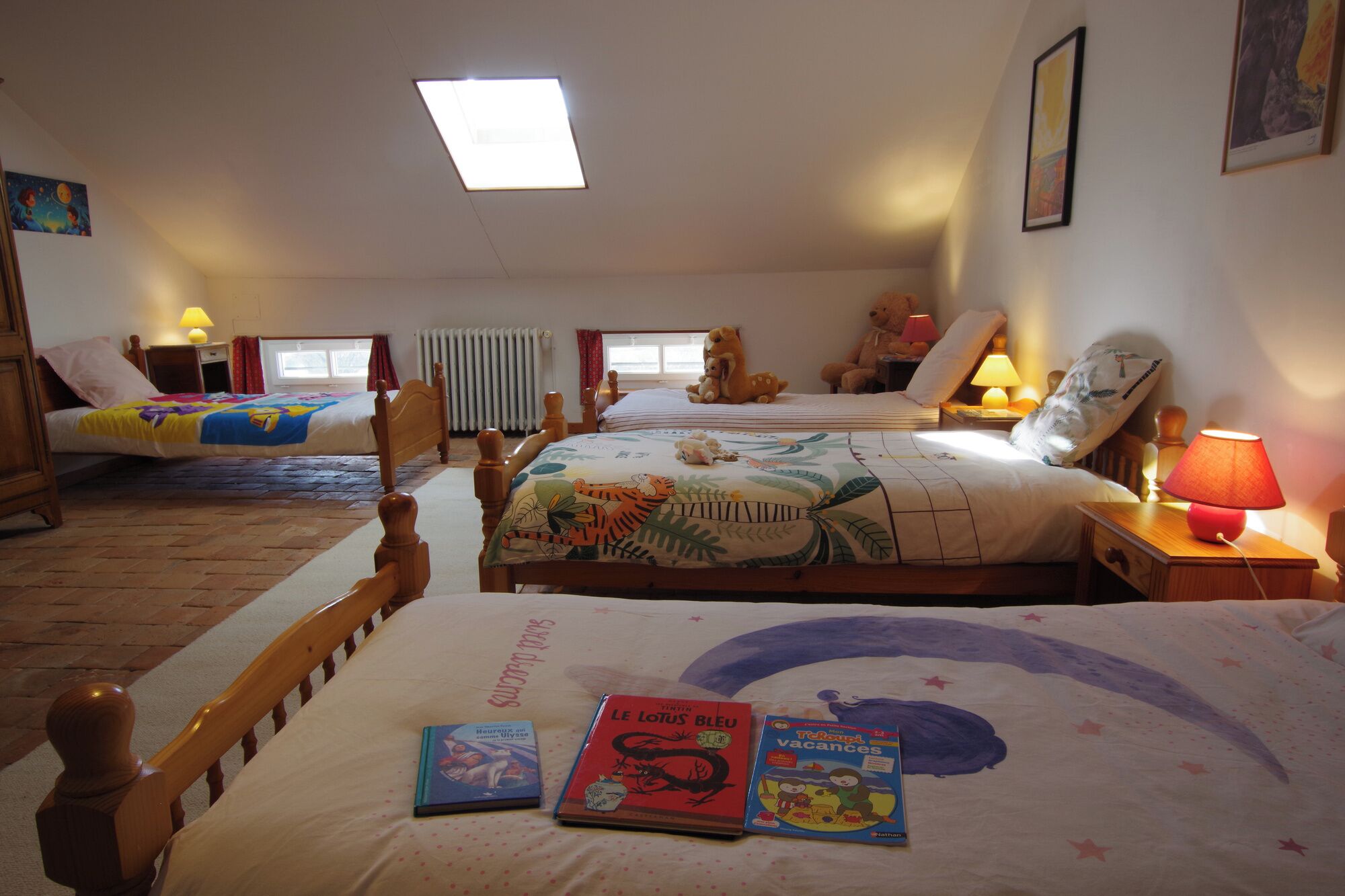 Une chambre confortable avec deux lits, une lucarne et une fenêtre. Il y a des livres sur les lits et un ours en peluche sur l’un d’eux.