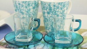 Two clear glass mugs with handles on matching saucers, placed on a table with a floral pattern.