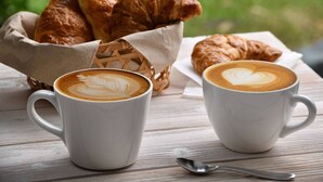 Two white cups of coffee with latte art on a wooden table, a basket of croissants, and a spoon.