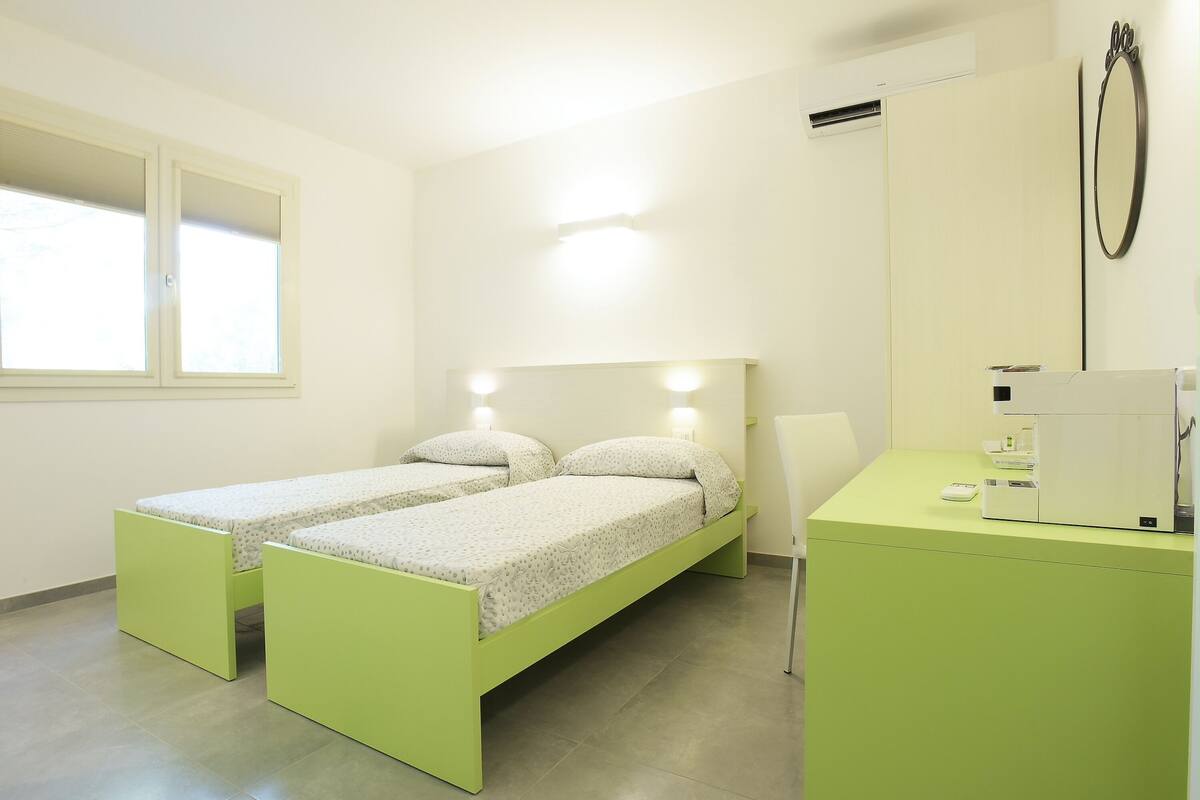 A room with two single beds, a desk with a computer, a mirror, and a window.
