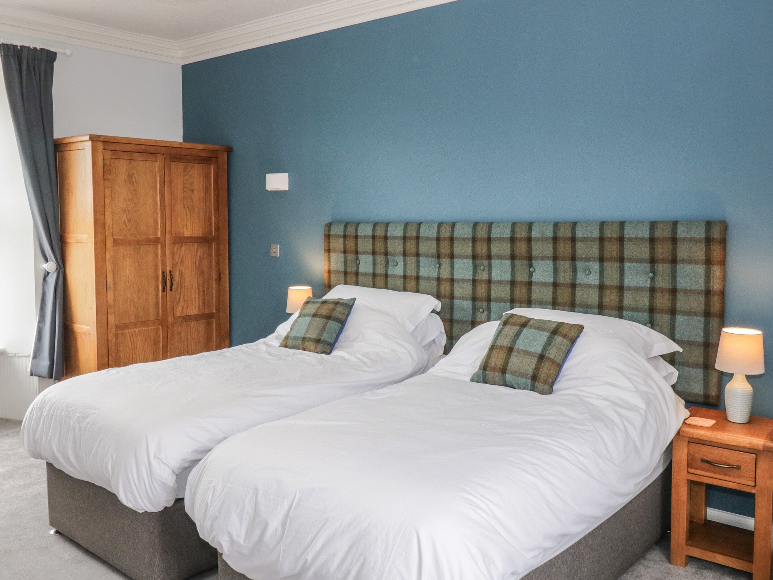A double bed with white bedding and plaid headboard, a wooden wardrobe, and a bedside table with a lamp.