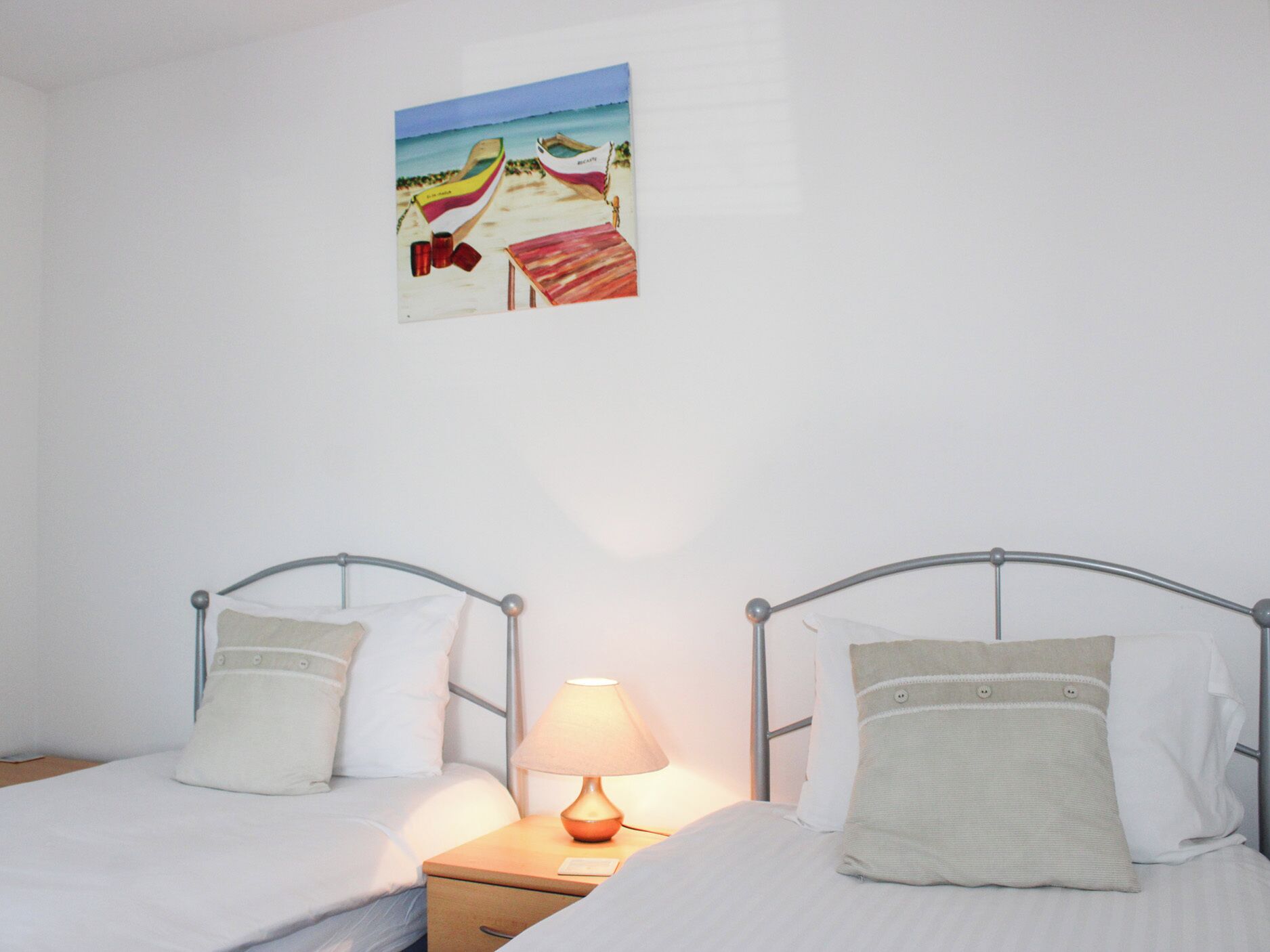 A bedroom with two beds, a nightstand with a lamp, and a beach-themed painting on the wall.