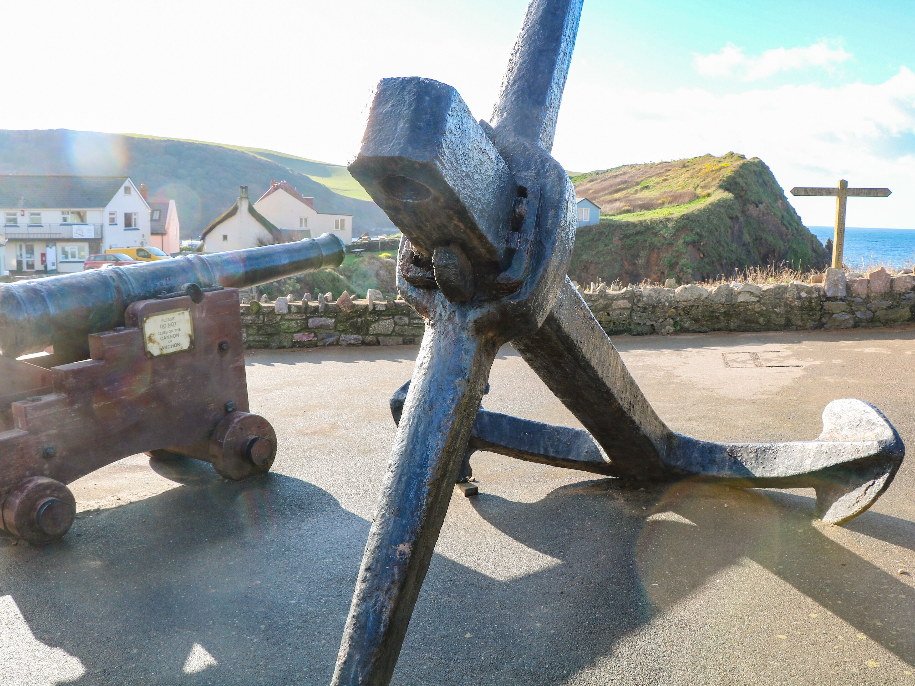 A cannon and anchor on a paved area with a stone wall and a hill in the background.