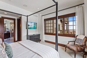 A bedroom with a four-poster bed, a large window with a view, a television on the wall, and a chair with a leopard print cover.
