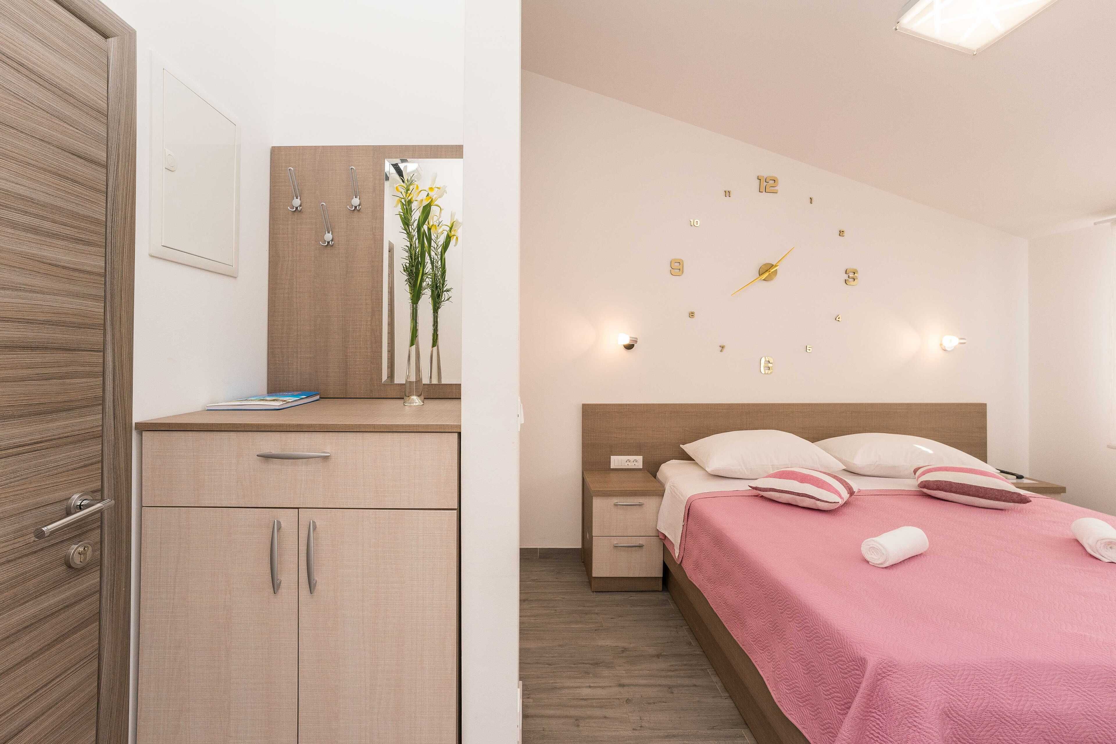 A modern bedroom with a wooden wardrobe, a bedside table with a vase of flowers, a bed with a pink bedspread, and a wall clock.